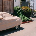 Car covered with a protective cover parked near a wooden fence and greenery